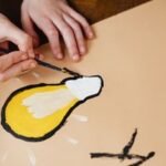 A person paints a yellow lightbulb design on paper with a paintbrush, symbolizing creativity.
